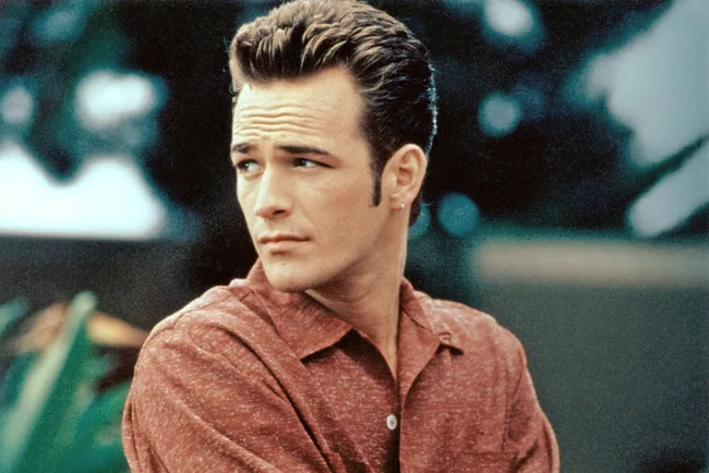 A man with styled hair in a red shirt looks to the side outdoors.
