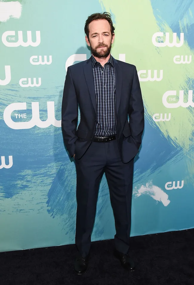 Man in a blue suit with a patterned shirt stands in front of a "The CW" backdrop.