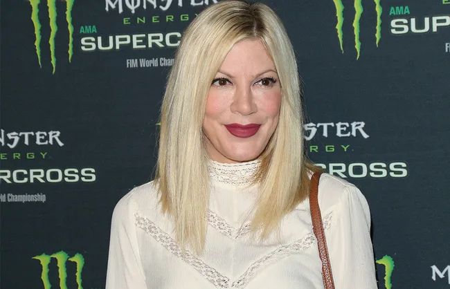 A person with long blonde hair wearing a white top, standing in front of a Monster Energy Supercross backdrop.