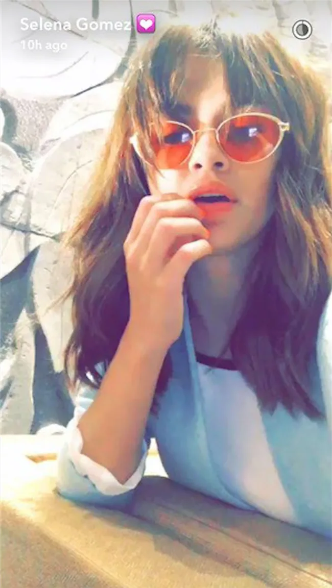 A woman with wavy hair and red sunglasses takes a selfie, hand near her mouth, wearing a light blue top.