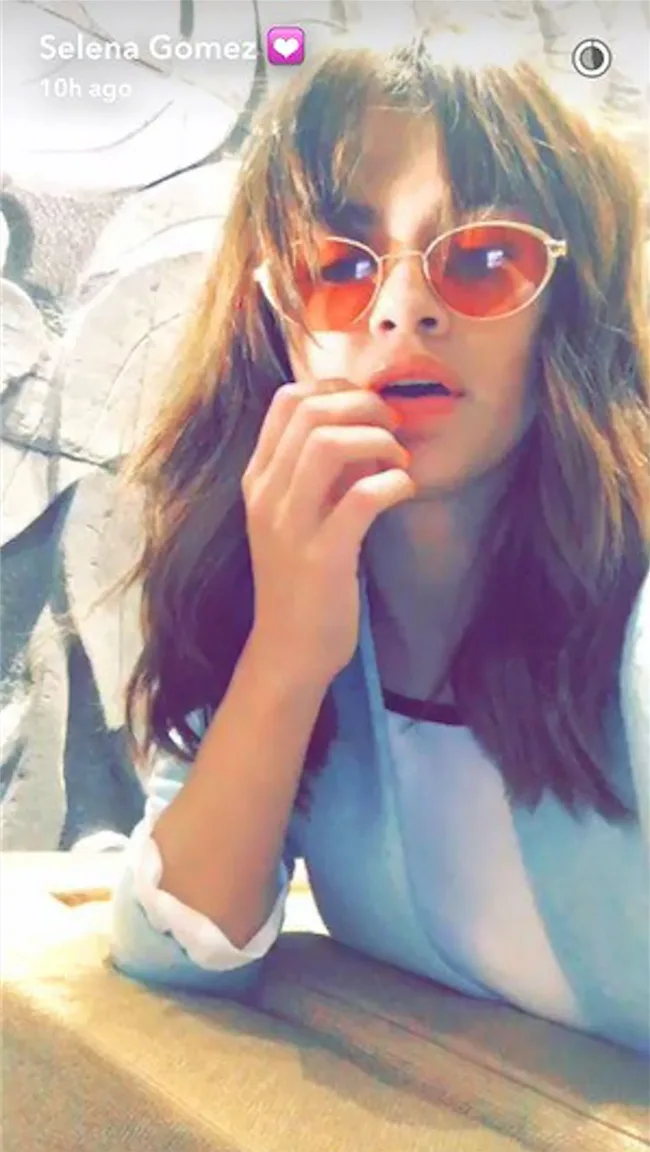 A woman with wavy hair and red sunglasses takes a selfie, hand near her mouth, wearing a light blue top.