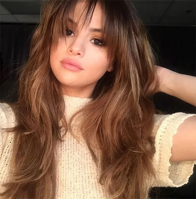 A person with long brown hair and bangs wearing a beige knit sweater poses with hand in hair, neutral expression.