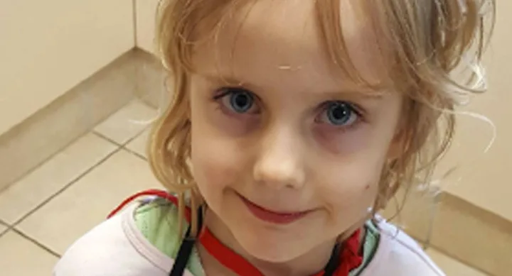 Child with blond hair and blue eyes smiling indoors.