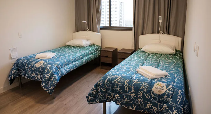 Two single beds with Rio 2016 themed bedspreads in a room, with a window and nightstands between them.