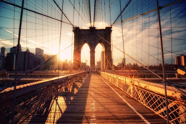 Brooklyn Bridge walkway at sunset with sun rays illuminating the structure and view of city skyline in background.