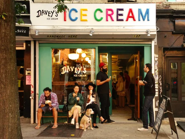 "People eating ice cream outside Davey's Ice Cream shop with colorful sign, tree in foreground."