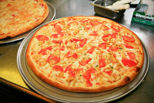 Pizza with sliced tomatoes, chicken, and cheese on a tray, with a plain cheese pizza in the background.