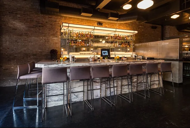 Modern bar interior with marble countertop, high chairs, and a well-stocked back shelf in a dimly lit setting.