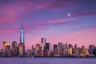 New York City skyline at dusk with One World Trade Center, under a purple and pink sky with the moon visible.