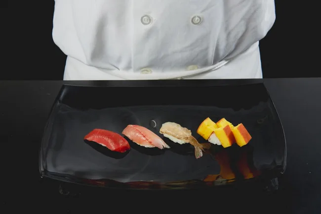 Sushi selection of tuna, salmon, prawn, and tamago on black plate, with chef in white coat standing behind.