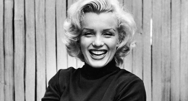 A person with short blond hair smiles brightly, wearing a dark top, with a wooden fence in the background.