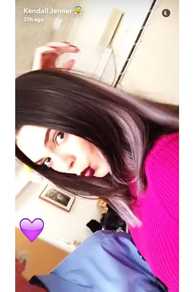 Young woman with dark hair and purple lipstick in a pink sweater, beside a purple heart emoji.