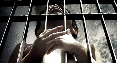 A person behind bars, looking up, with hands grasping the prison cell in a shadowy, dramatic setting.