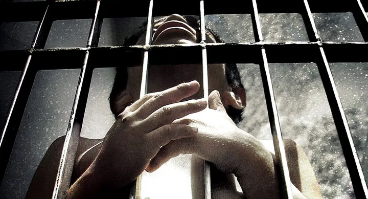 A person behind bars, looking up, with hands grasping the prison cell in a shadowy, dramatic setting.