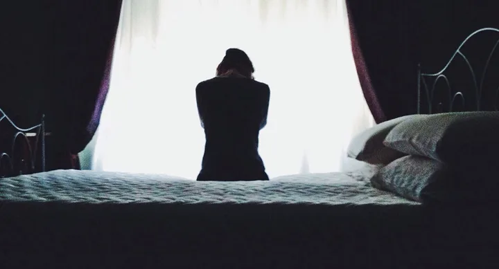 "Silhouette of a person sitting on a bed with curtains backlit by bright light."