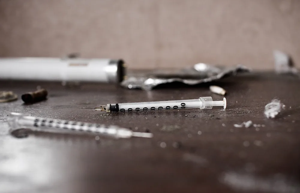 Syringes and drug paraphernalia scattered on a surface, with a blurred background.