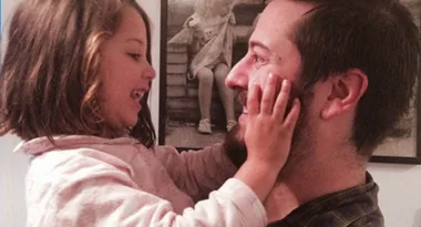 A child smiles, touching a man's face affectionately indoors, with black and white photos in the background.