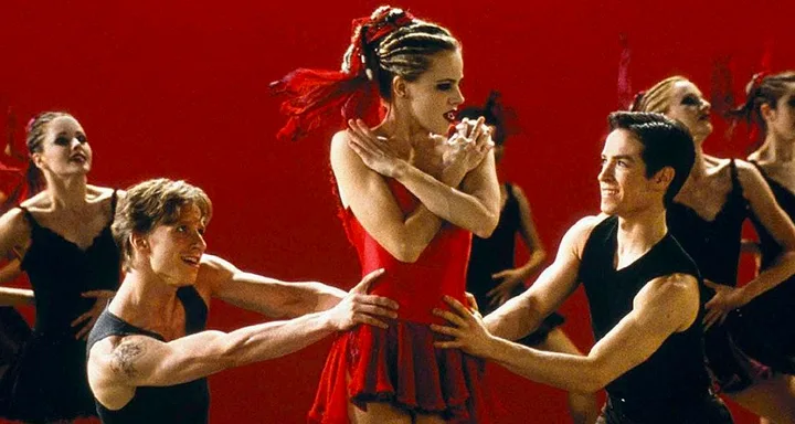 Dancers performing a ballet routine; a woman in a red dress is centered, surrounded by male and female dancers.