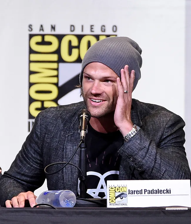 Actor in beanie speaks at San Diego Comic-Con panel, smiling with hand to face.