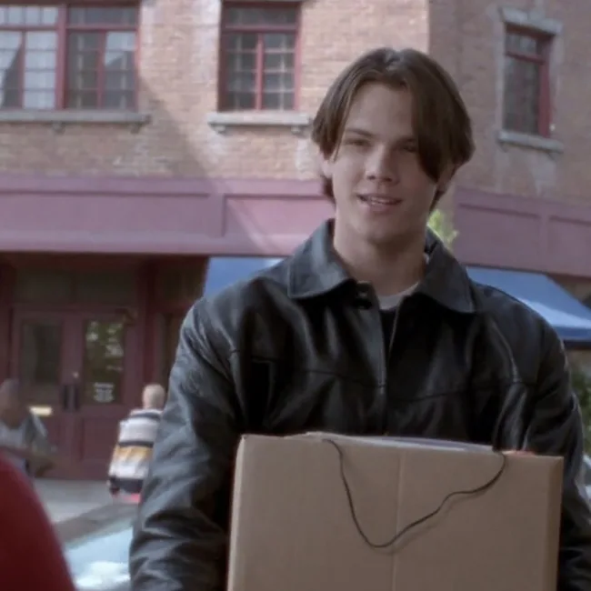 Young man with medium-length hair in a leather jacket holding a cardboard box outdoors.