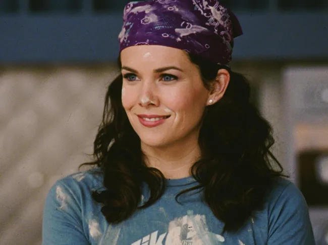 A woman with dark hair and a purple bandana smiles, wearing a blue shirt with white paint stains.