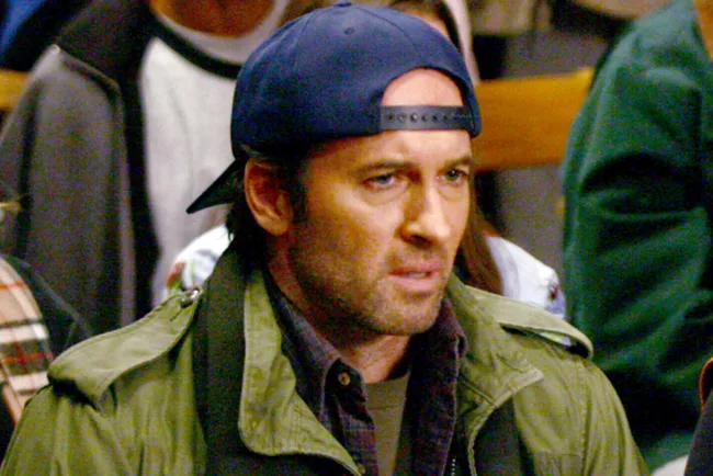 A man wearing a backward cap and green jacket looks focused in a crowded room.