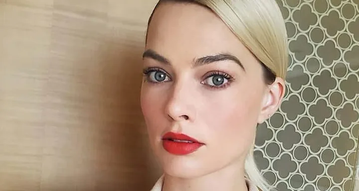 A woman with blond hair, red lipstick, and blue eyes, wearing a tan top, stands against a patterned wall.