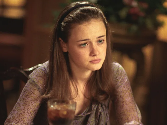 Young woman with long brown hair, wearing a purple-patterned top, sits at a table looking thoughtful.