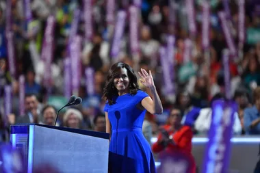 Woman in a blue dress waves on stage at a large event with an audience in the background.