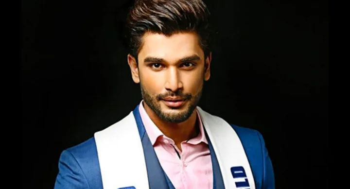 A man in a suit with a white sash poses confidently against a dark background.