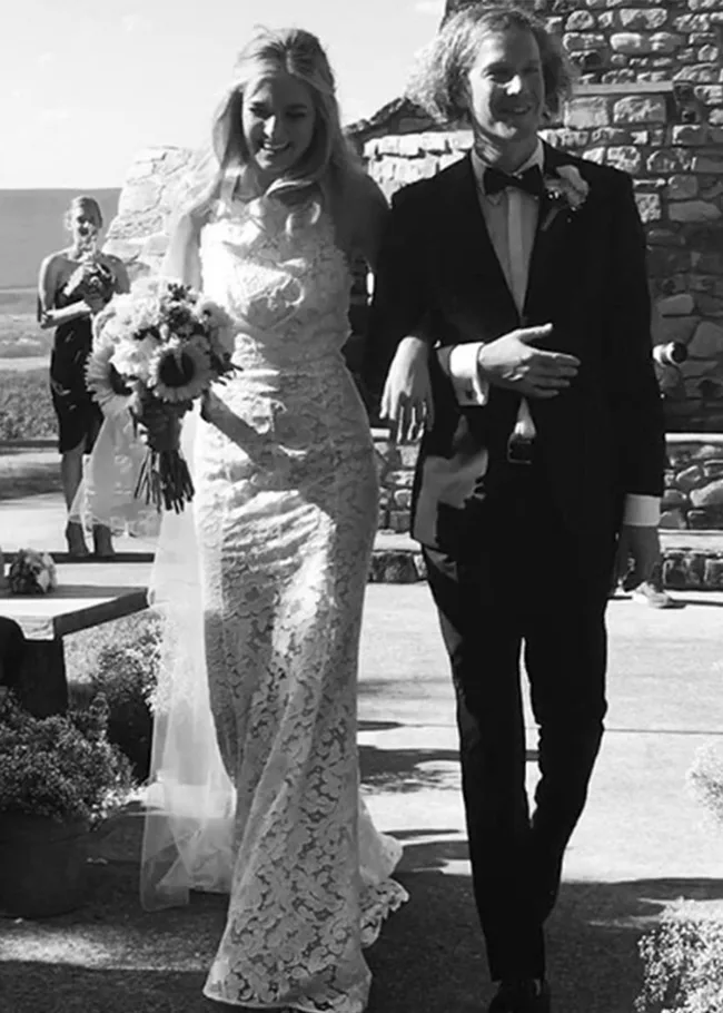 Bride in lace gown holding bouquet, escorted by a man in a tuxedo, smiling outdoors during a wedding ceremony.