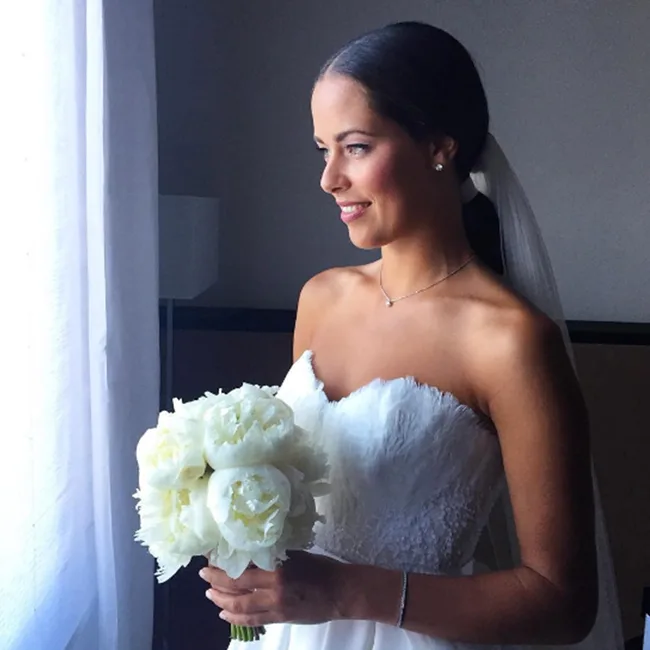 Bride in strapless white gown holding a bouquet, looking out of a window with a gentle smile.