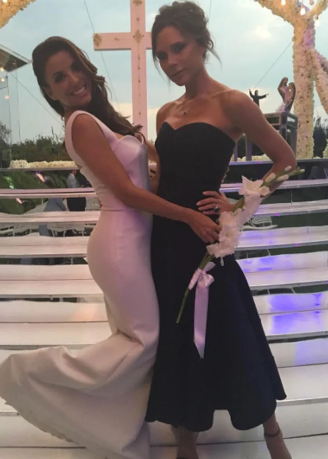Two women pose elegantly, one in a white dress and the other in black, in front of a decorated cross at an event.