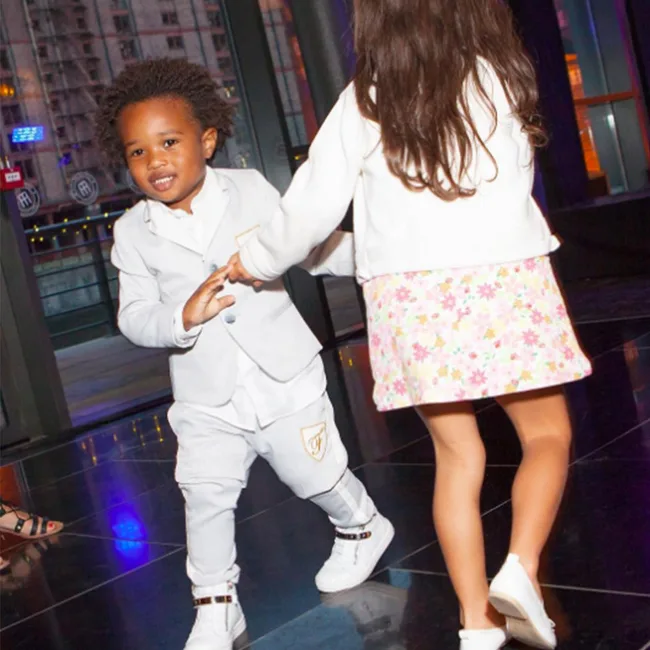 Two children dancing indoors, the boy in a gray suit and the girl in a white top with a floral skirt.