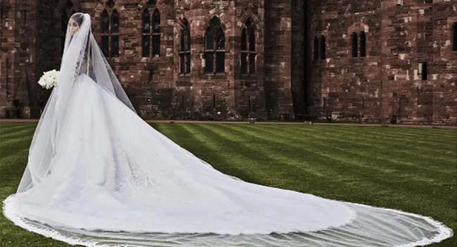Bride in an elegant white gown with a long train, standing on lush green grass in front of a historic stone building.