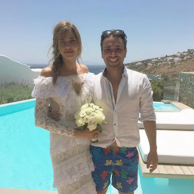 Couple by a pool, woman in a lace dress holding flowers, man in colorful shorts, with a coastal background.