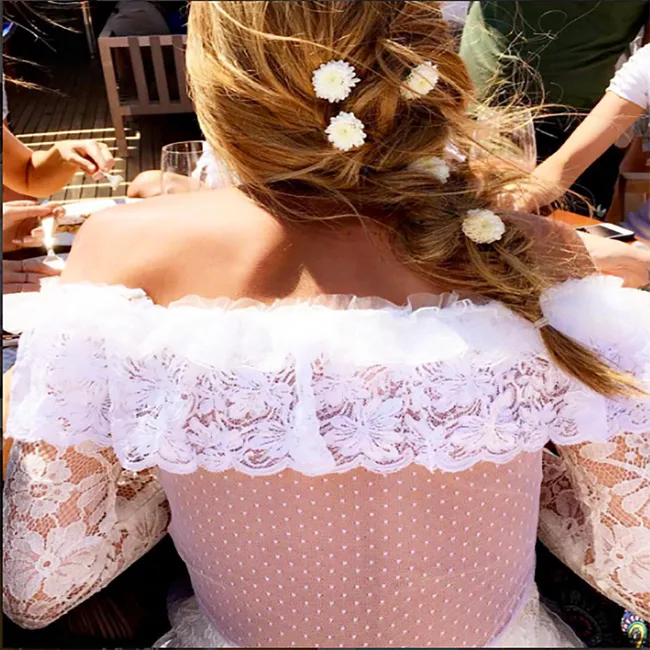 Woman in lace off-shoulder dress with flowers in braided hair, sitting outdoors with others.
