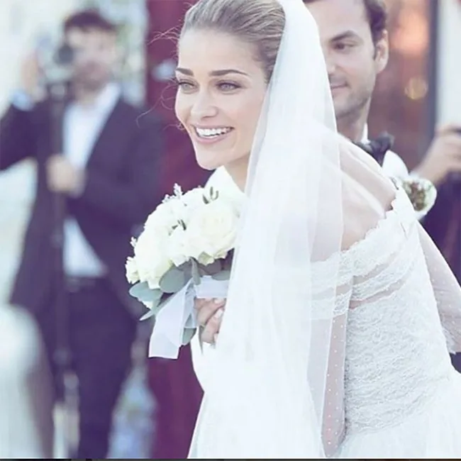 Bride smiling in an off-shoulder lace gown, holding a white bouquet, with a man and photographer in the background.