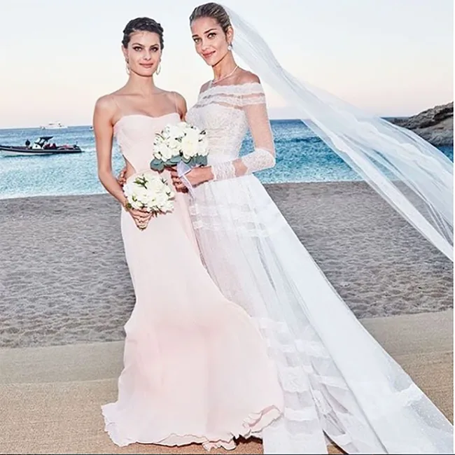 Two women on a beach, one in a wedding dress with veil, the other in a blush dress, holding bouquets, ocean in background.
