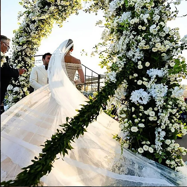 Bride in flowing gown stands under floral arch at outdoor wedding ceremony.