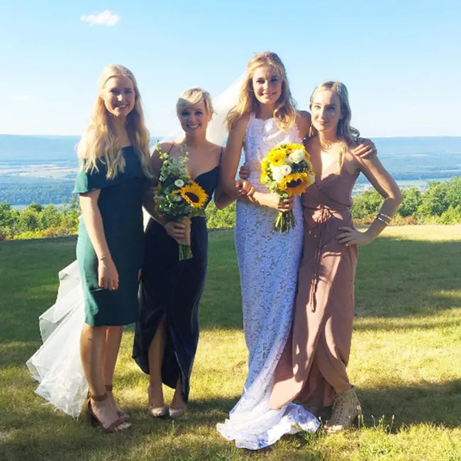 Four women in dresses, two holding bouquets with sunflowers, pose outdoors with a scenic view in the background.