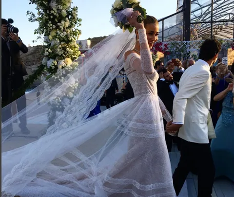 Bride in lace gown holding bouquet, walking with groom in white jacket at outdoor wedding ceremony.