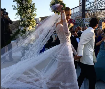 Bride in lace gown holding bouquet, walking with groom in white jacket at outdoor wedding ceremony.