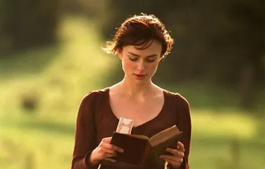 A woman in period clothing reads a book outdoors with a green, blurred background.