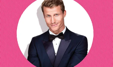 Man in a dark tuxedo and bow tie smiling against a pink and white striped background.