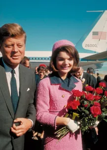 Man in suit and woman in pink outfit holding roses, standing outside near an airplane.