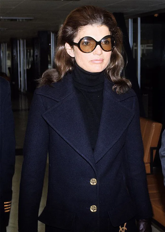 A woman in a dark coat and large sunglasses indoors, with wavy hair, looking ahead.