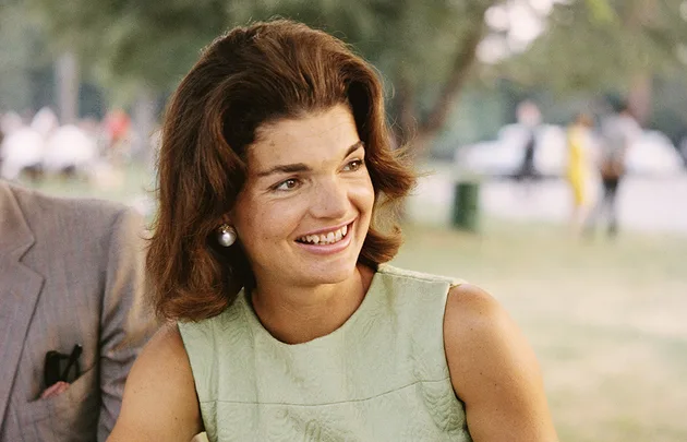 A woman with brown hair and pearl earrings smiles outdoors, wearing a light green sleeveless top.