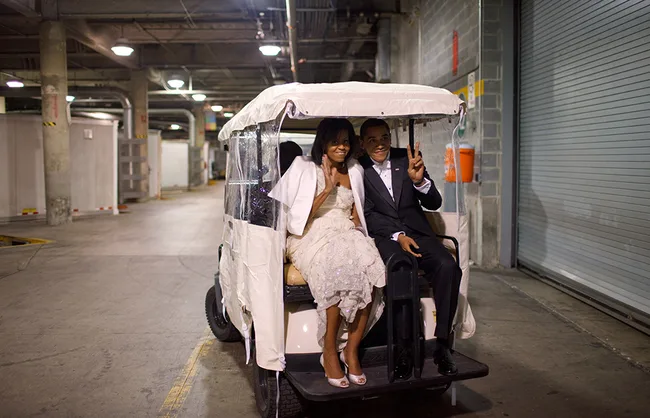 Two people in formal attire sit on a golf cart in an underground parking area, both making peace signs.