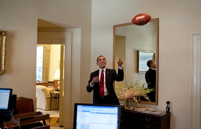 Man in a suit tossing a football inside an office, reflected in a mirror; computers and floral arrangement visible.